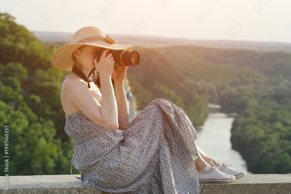 Girl in a sundress and a hat with a camera. Green forest and river in the distance
