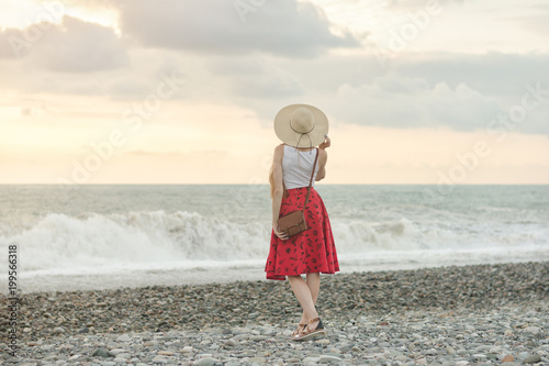 Girl in red skirt and hat standing by the sea, sunset. Back view