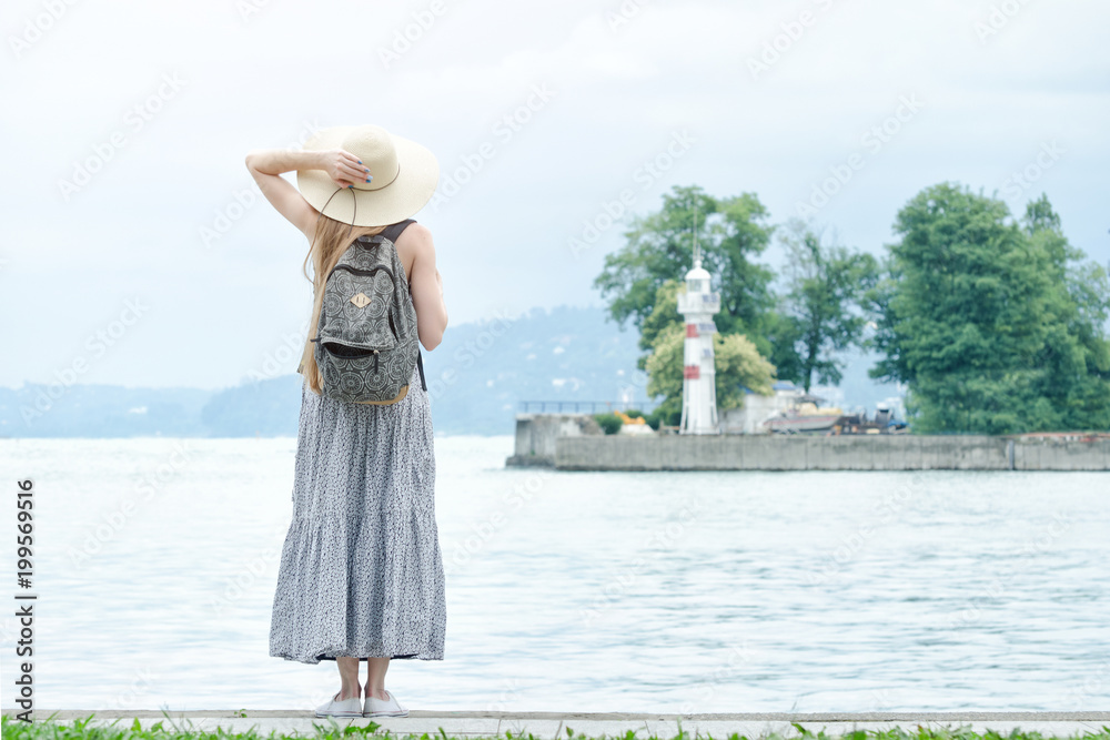 Girl in a hat with a backpack standing on a pier at the sea. Mountains and lighthouse on the background. Back view