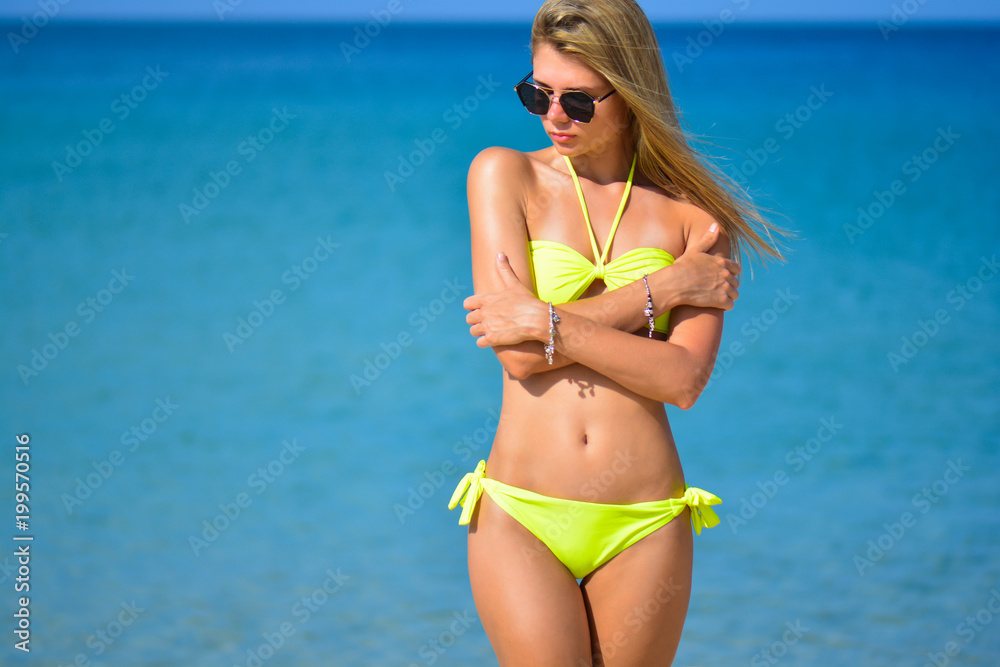 beautiful fit young woman in sexy yellow bikini at the beach. Girl in swimsuit and sunglasses, summer time near blue sea