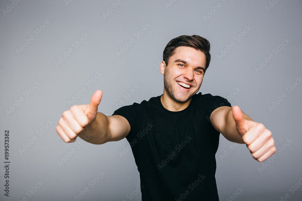 Fototapeta premium Happy successful confident man in black t-shirt showing thumbs up . Handsome young man gesturing and keeping mouth open while standing against grey background