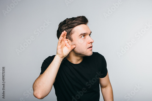 Curious handsome young man in shirt overhearing rumors over grey background.