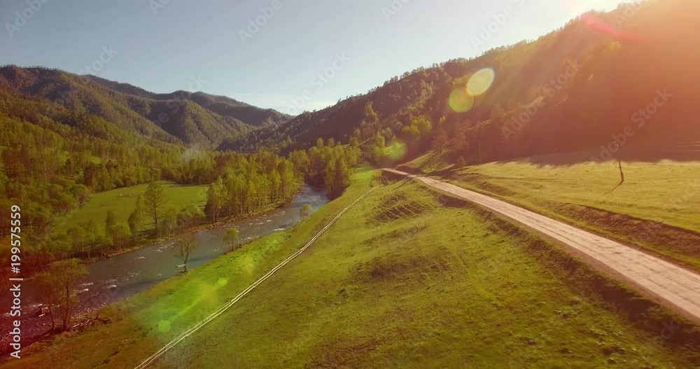 Aerial UHD 4K view. Mid-air flight over fresh mountain river and meadow at sunny summer morning. Green trees and sun rays on horizon. Rural dirt road below. Aerial UHD 4K view. Mid-air flight over fresh mountain river and meadow at sunny summer morning. Green trees and sun rays on horizon. Rural dirt road below.