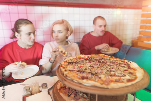 A delicious large pizza on the background of people. A group of young people sits in a cozy pizzeria and eats a delicious pizza