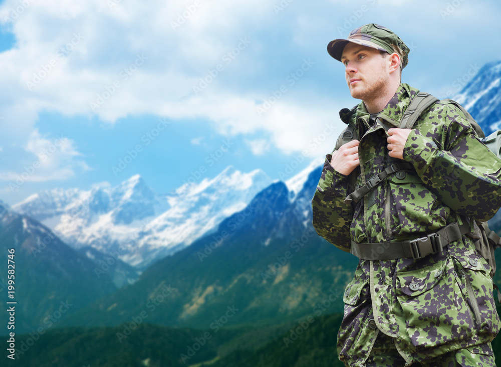 army, military service and people concept - young soldier or traveler in camouflage uniform with backpack hiking over mountains background