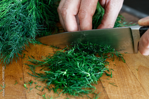 Detail of hands chopping up a bunch of dill on wood chopping board with a kitchen knife.