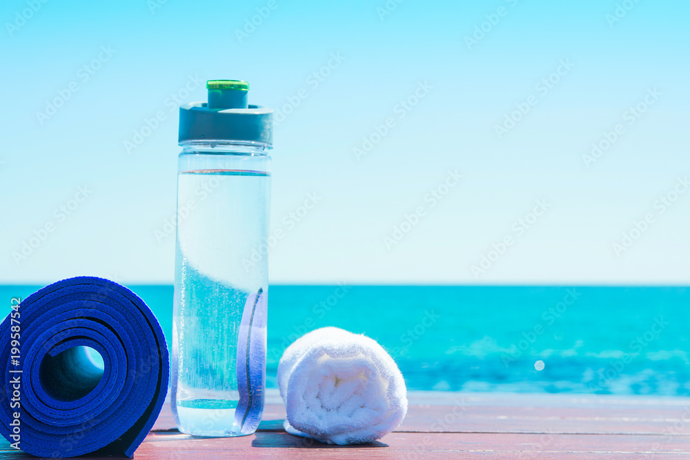 Rolled Yoga Mat Bottle with Water White Towel on Beach with Turquoise Sea  Blue Sky in Background. Sunlight. Relaxation Summer Meditation Fitness  Wellbeing Mindful Living Concept. Copy Space Stock Photo