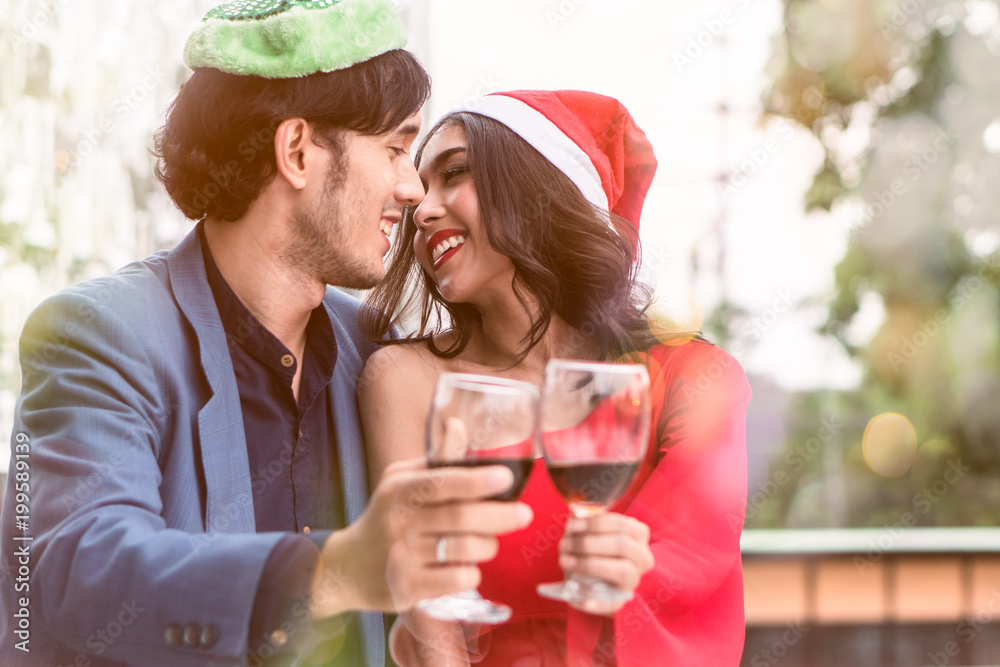 Lovely couple romantic christmas time. Young couple kissing in the christmas eve party and holding wine glass in hands. decorated with green fancy hat and red santa hat on the lovers. Lovely couple romantic christmas time. Young couple kissing in the christmas eve party and holding wine glass in hands. decorated with green fancy hat and red santa hat on the lovers.