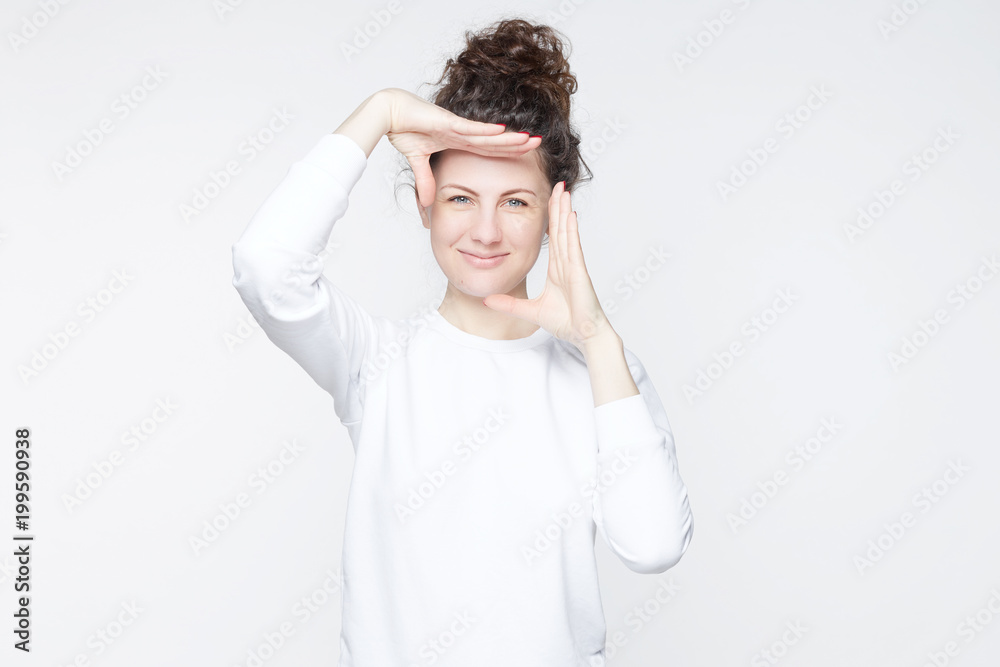Indoor studio portrait of beautiful young Caucasian woman with hair bun looking at camera and gesturing finger frame, while standing against isolated white wall background. Body language concept. Indoor studio portrait of beautiful young Caucasian woman with hair bun looking at camera and gesturing finger frame, while standing against isolated white wall background. Body language concept.
