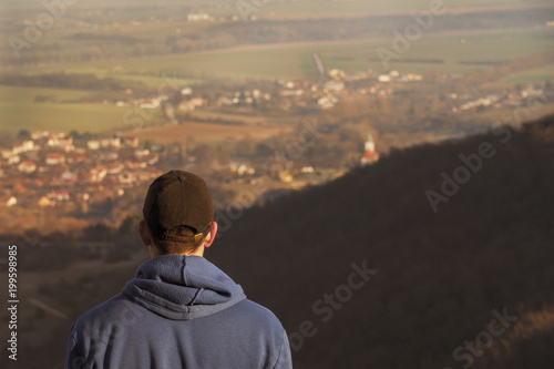 Adult man (hiker) standing on the rock and enjoying view with blurred village on background. Silhouette of tourist man in forest at sunset.