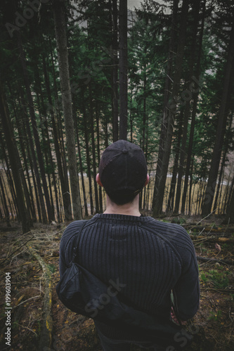 Young caucasian man (hiker) standing in the forest and enjoying perfect view on trees. Man relaxing and chilling in the forest (image from his back)