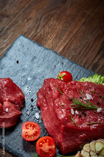 Raw meat beef steaks on black slate board with spices, garlic and rosemary over wooden background, selective focus
