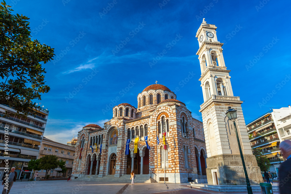 Stone Greek Orthodox Church on the town square with the city clock on the tower, among modern houses, summer, blue clear sky. Cathedral Church of St. Nicholas, Volos, Greece - April 2017.