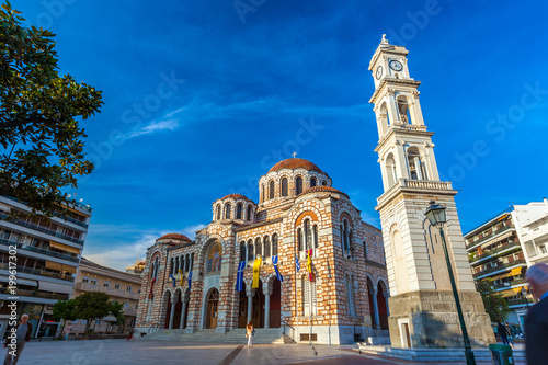 Stone Greek Orthodox Church on the town square with the city clock on the tower, among modern houses, summer, blue clear sky. Cathedral Church of St. Nicholas, Volos, Greece - April 2017.