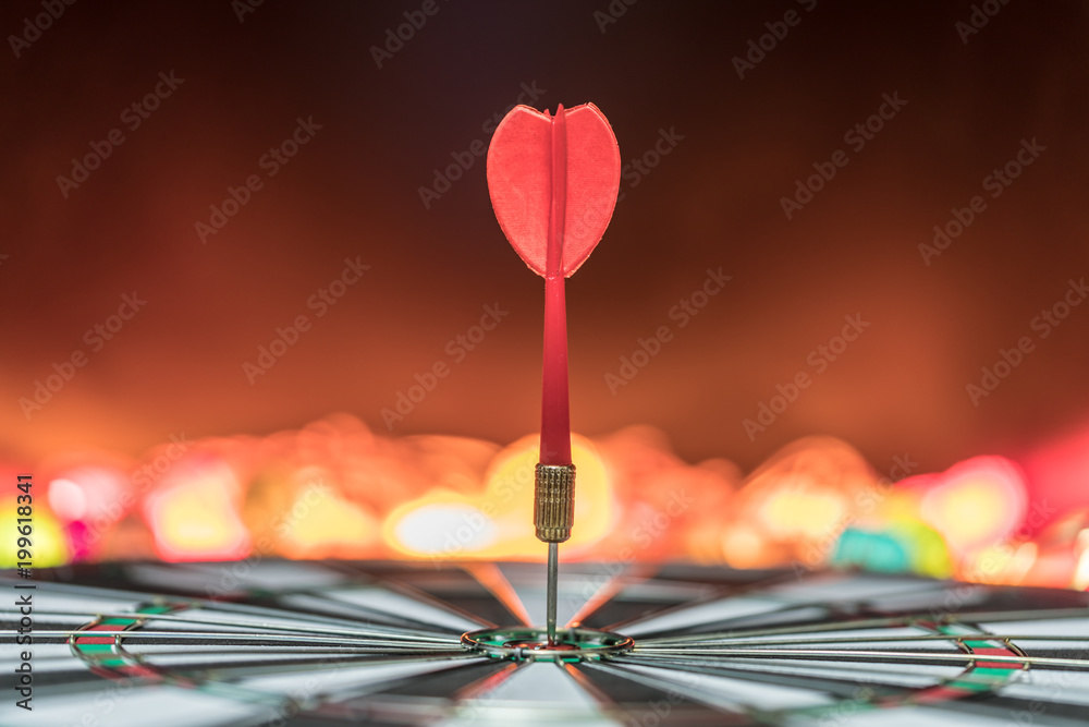 Red dart arrow hitting in the target center of dartboard with colorful bokeh and black background. Target business, achieve and victory concept . Red dart arrow hitting in the target center of dartboard with colorful bokeh and black background. Target business, achieve and victory concept .