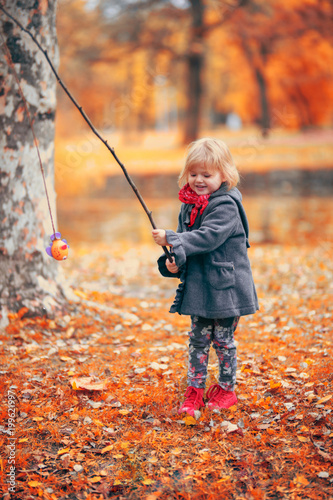 Beautiful little girl playing fishing with a branch and fish toy, in the park on a cold autumn day