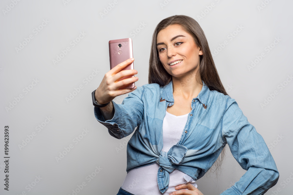 Portrait of a young smiling brunette woman in casual clothes making a selfie with her pink phone against white background. Lifestyle, people and technology concept Portrait of a young smiling brunette woman in casual clothes making a selfie with her pink phone against white background. Lifestyle, people and technology concept
