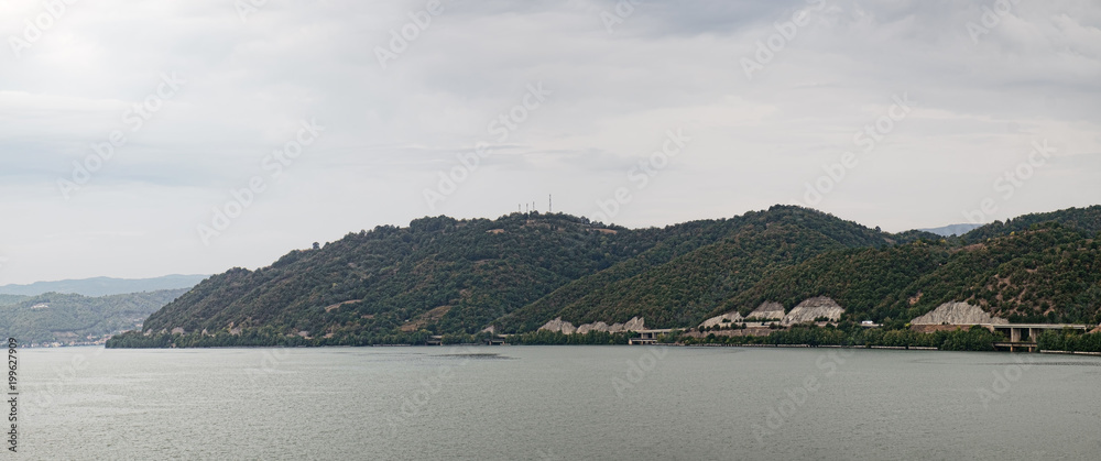 A picturesque landscape with a river bank covered with forest and road along the shore. Panoramic view of the point where the Cerna River meets the Danube, southwestern Romania's Mehedinti County.