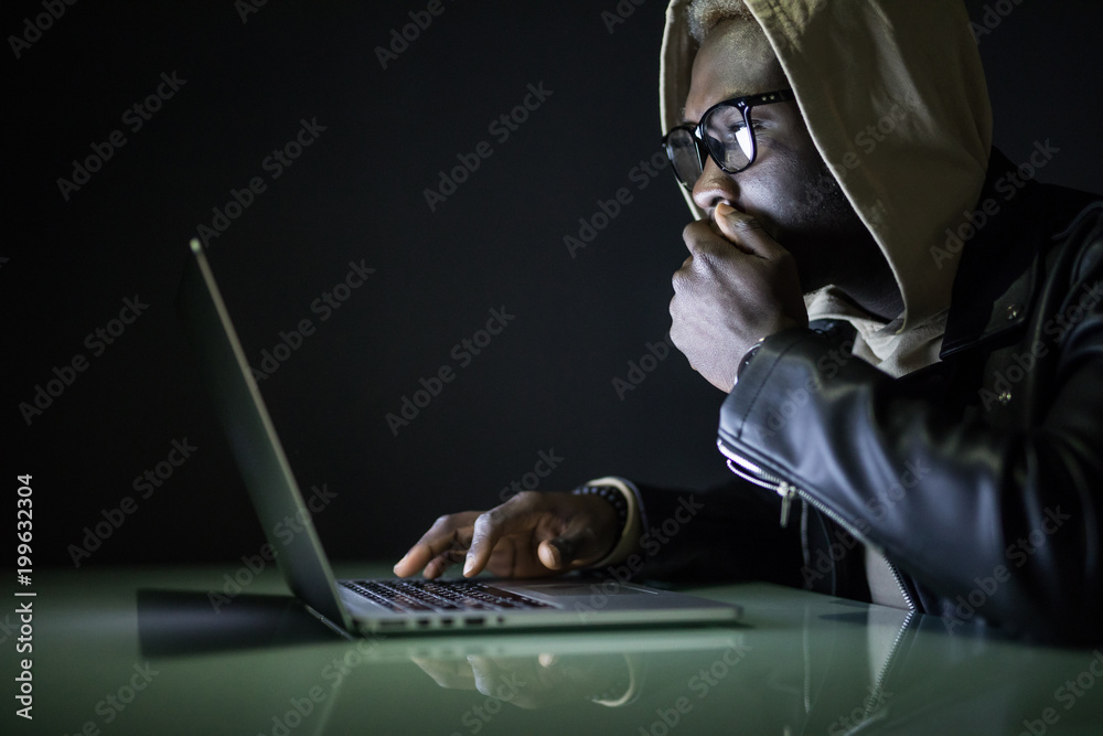 Afro american man sits at night at a computer desk using the Internet. Night surfing on the Internet. Gamer sits at night at a table near a computer with cookies in his hands and looks at the monitor Afro american man sits at night at a computer desk using the Internet. Night surfing on the Internet. Gamer sits at night at a table near a computer with cookies in his hands and looks at the monitor