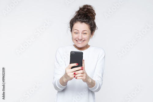 Excited female model receives unexpected message or news on telephone, looks joyfully into gadget, has overjoyed expression. Positive young happy woman views funny instagram photos on mobile phone.
