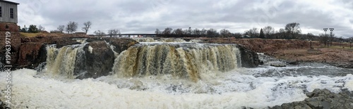 The Big Sioux River flows over rocks in Sioux Falls South Dakota with views of wildlife, ruins, park paths, train track bridge, trees and city in the surrounding area and background