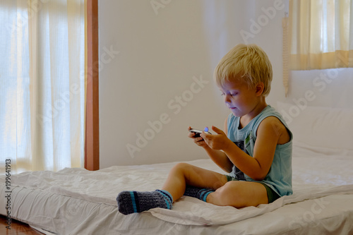 Little cute boy enthusiastically playing video games on portable device. For fun and joy. Sitting on mattress. Asian domestic room interior