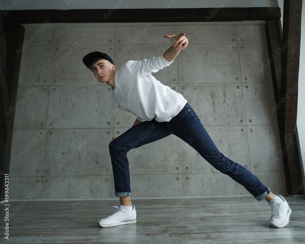 Teenage boy in white sweater and baseball cap, jeans and sneakers dances hip-hop in dance studio. Dynamics of modern dance movement.