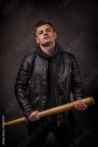 The young man leather jacket on a black wall in fighting pose with baseball bat