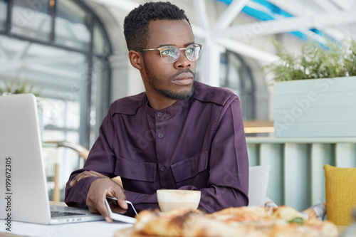 Pensive dark skinned African male freelancer in elegant shirt, works on new project, holds modern smart phone, drinks coffee, finds some important information. Serious businessman has thoughtful look