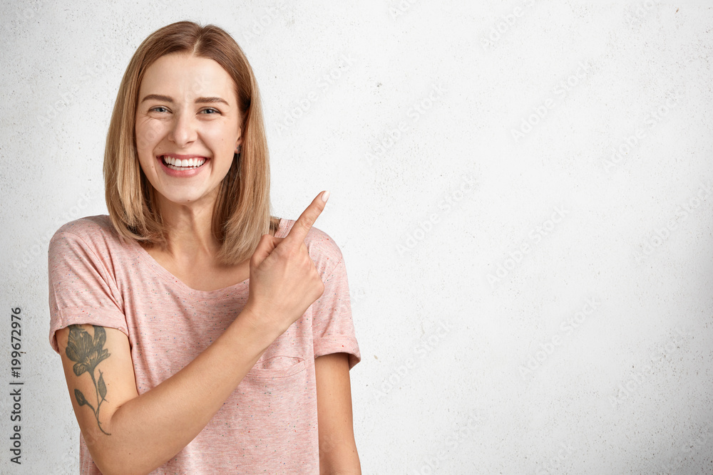 Beautiful young female with positive expression has tattoo, smiles pleasantly and indicates at blank copy space for your promotional text or advertising content, isolated over white concrete wall Beautiful young female with positive expression has tattoo, smiles pleasantly and indicates at blank copy space for your promotional text or advertising content, isolated over white concrete wall