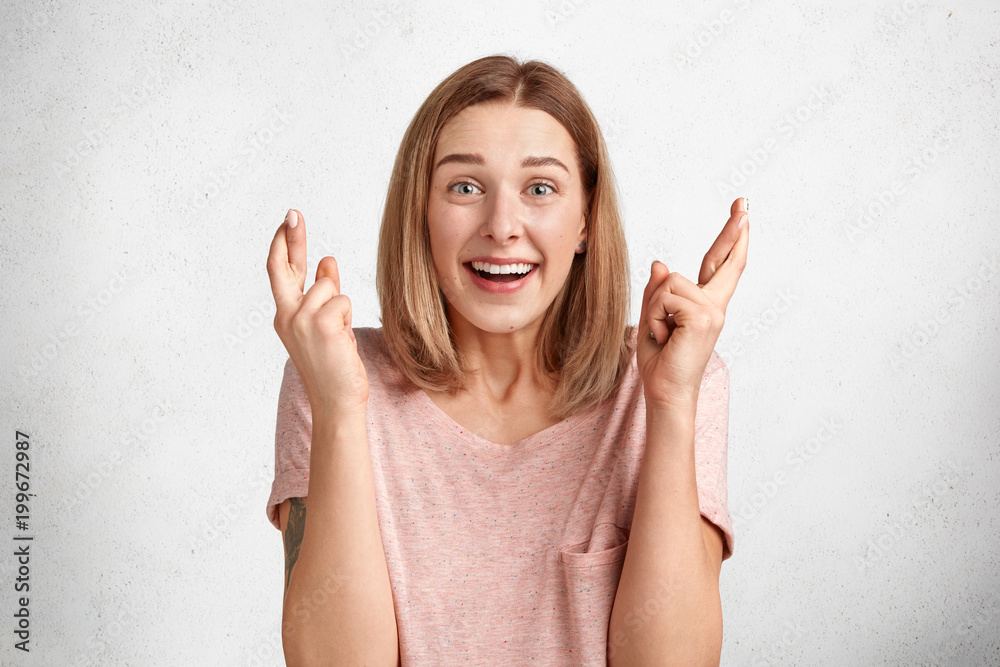 Overjoyed young Caucasian female with pleasant apperance and broad smile, crosses fingers as makes desired wish, dressed in casual t shirt, poses against white concrete background. Believe in success Overjoyed young Caucasian female with pleasant apperance and broad smile, crosses fingers as makes desired wish, dressed in casual t shirt, poses against white concrete background. Believe in success