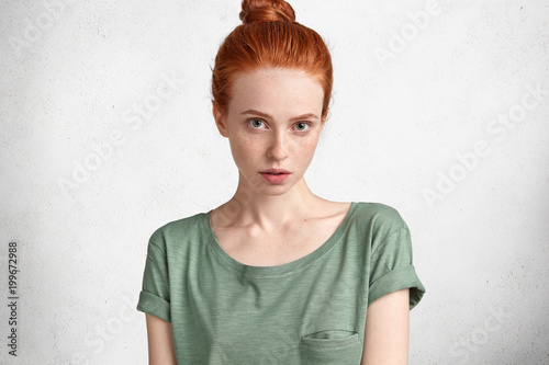 Studio shot of beautiful red haired woman with healthy freckled skin, dressed casually, has serious expression, isolated over white concrete wall. Gorgeous ginger teenage girl poses in studio