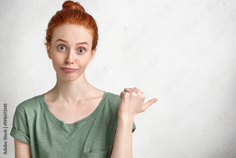 Horizontal portrait of puzzled ginger femalee with hair knot, dressed in casual t shirt and has clueless look, indicates at blank copy space for your promotional text or advertizing background Horizontal portrait of puzzled ginger femalee with hair knot, dressed in casual t shirt and has clueless look, indicates at blank copy space for your promotional text or advertizing background