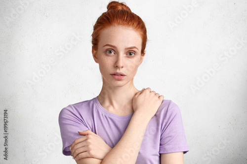 People, facial expressions and emotions concept. Adorable freckled red haired female model has hair knot, dressed in casual clothing, poses against white concrete wall with serious expression