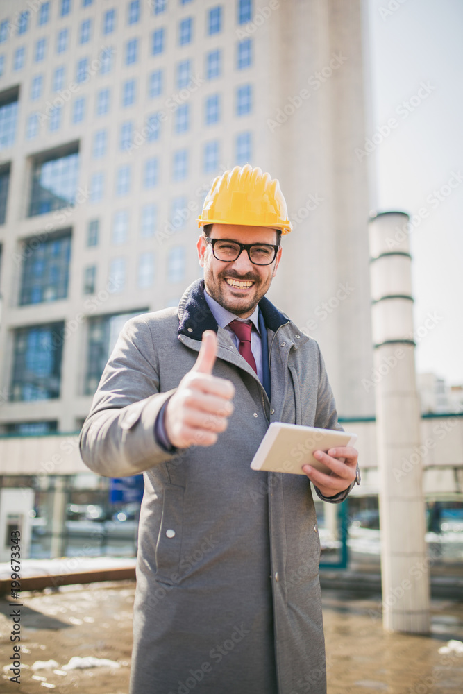 Architect in protective helmet showing thumb up in front of office building, holding digital tablet. Architect in protective helmet showing thumb up in front of office building, holding digital tablet.