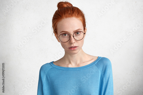 Indoor shot of confident serious female student wears glasses, has red hair bun, listens attentively what information she should prepare, isolated over white concrete background. Facial expressions