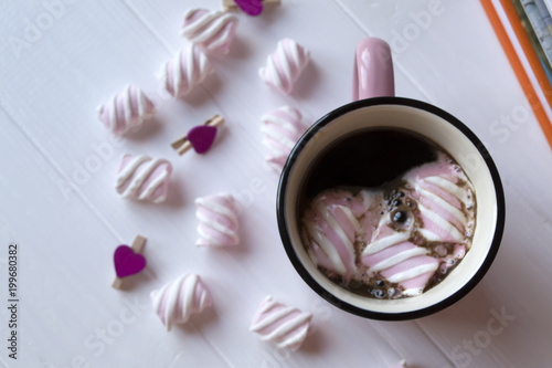 The pink mug of hot coffee with marshmallow on a white wooden table.