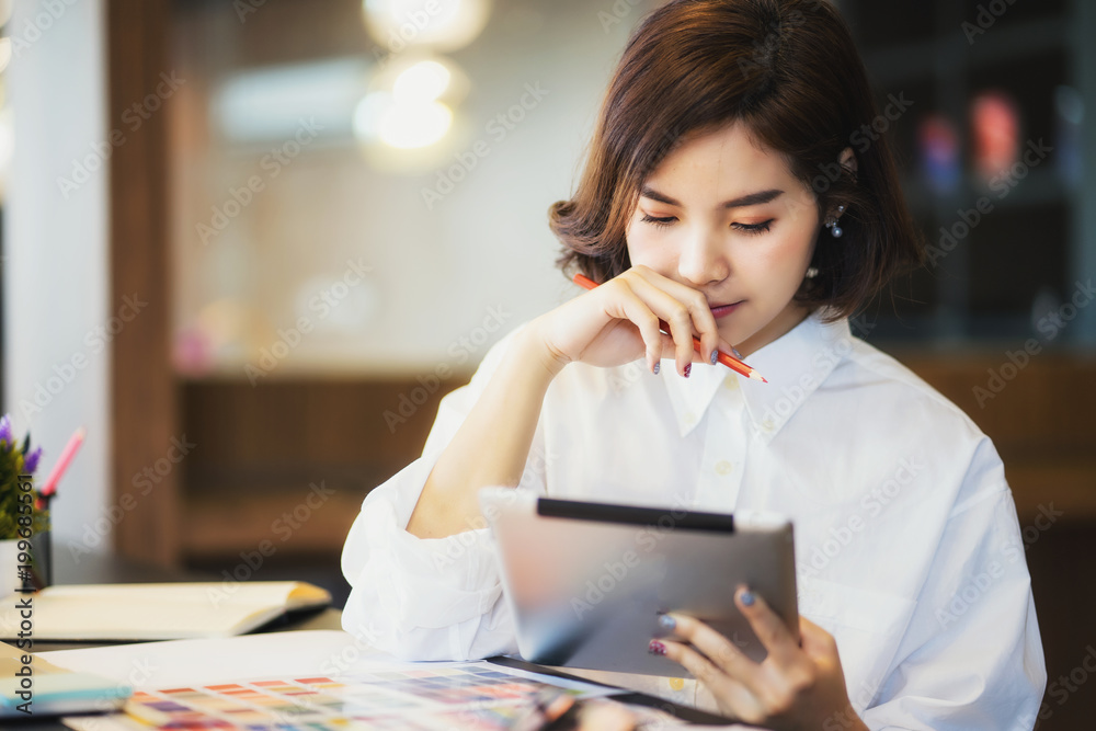 Young beautiful asian woman in white shirt holding pencil while looking at tablet Young beautiful asian woman in white shirt holding pencil while looking at tablet