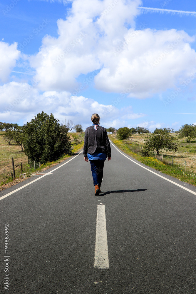 Young women walking alone in a Road with green grass field under white clouds and blue sky. Country side landscape in Portugal Young women walking alone in a Road with green grass field under white clouds and blue sky. Country side landscape in Portugal