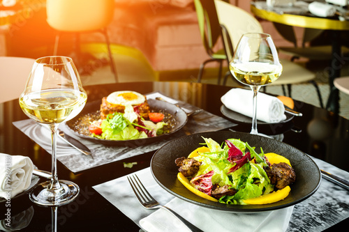 Romantic dinner at a restaurant. Appetizing dishes with meat and lettuce leaves and glasses with wine in rays of the sun