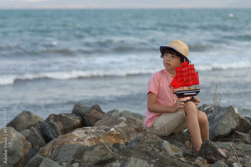 The boy sits on a stony seashore, holds a toy sailboat in his hands and looks into the distance.