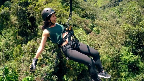 A woman in safety gear ziplines through a dense green tropical forest near Banos, Ecuador, arms outstretched, a high-angle view capturing her movement above the treetops.
