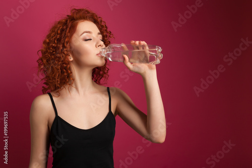 Pretty healthy young woman drinking pure mineral bottled water on a pink background