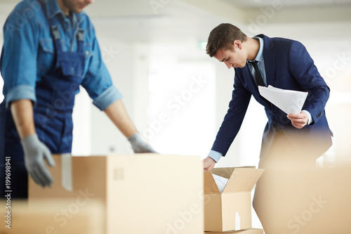 Contemporary businessman in suit looking for necessary papers in one of boxes just after relocation to new office