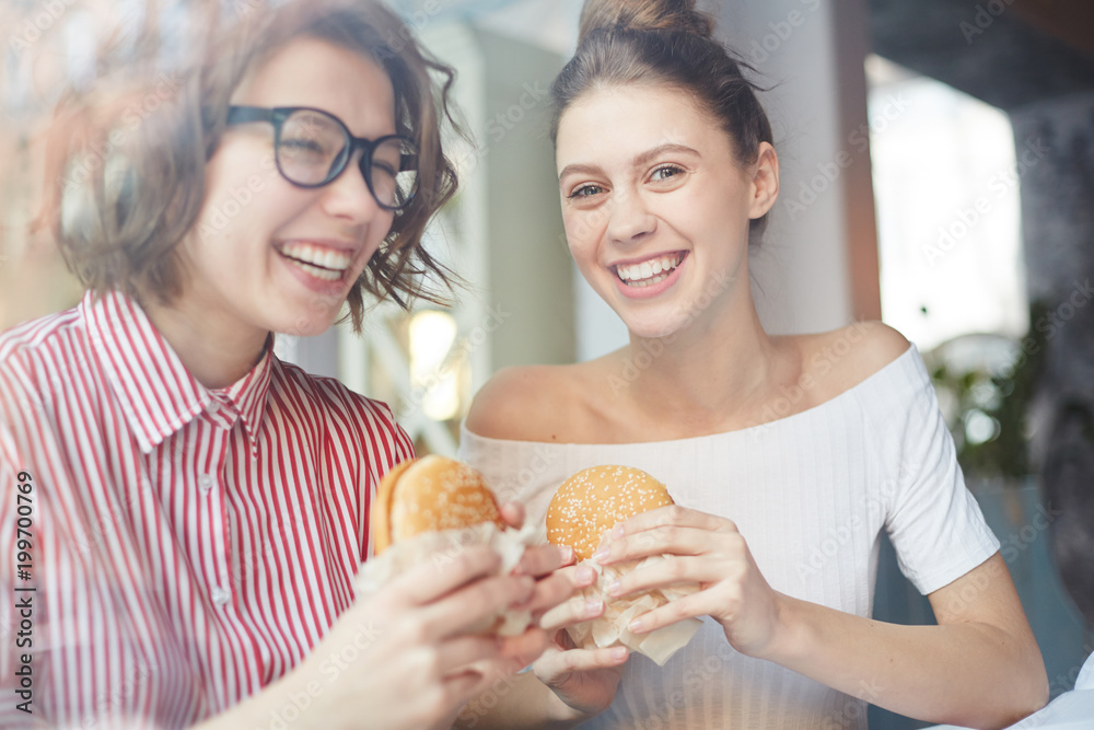 Two happy girls with appetizing and fat cheeseburgers having fun and enjoying time in fastfood cafe