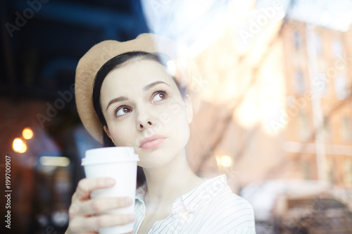 Pretty young woman in beige beret having drink in cafe and looking through window at urban scene