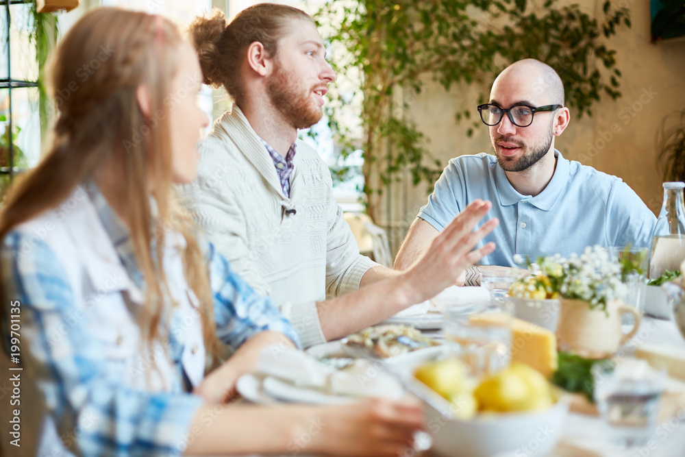 One of young friends telling something by served table during dinner or gathering in cafe One of young friends telling something by served table during dinner or gathering in cafe