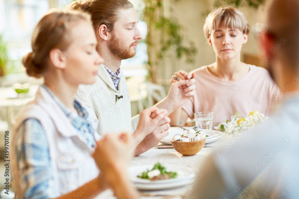 Group of friends sitting by served table and holding by hands while praying and blessing food before eating Group of friends sitting by served table and holding by hands while praying and blessing food before eating