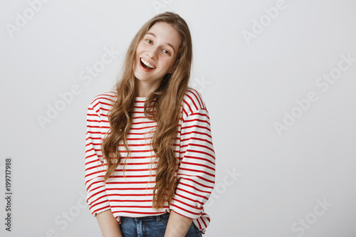 Positive teenager greets her best friends. Indoor shot of silly good-looking young woman standing in cute pose and smiling broadly while saying hi, being feminine and tender over gray background