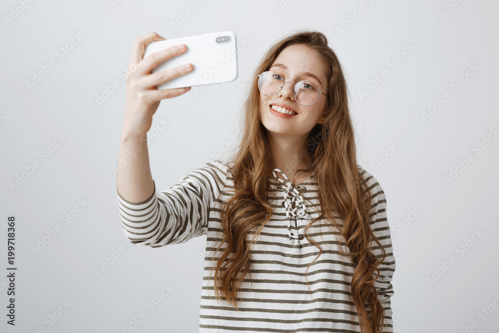 Modern teenagers cannot spend time without smartphones. Portrait of attractive urban girl taking selfie on phone, smiling and expressing positive emotions, standing over gray background Modern teenagers cannot spend time without smartphones. Portrait of attractive urban girl taking selfie on phone, smiling and expressing positive emotions, standing over gray background